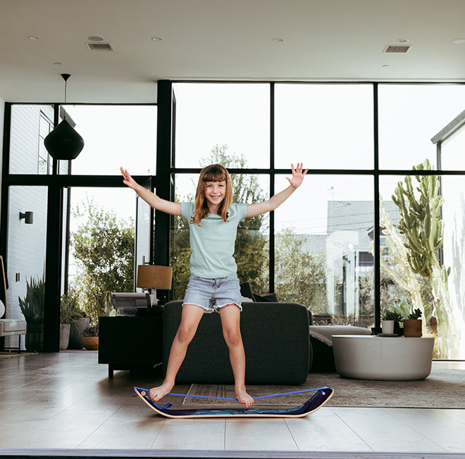 Young girl balancing on a Butterfly SlackBoard in a modern living room with large windows.