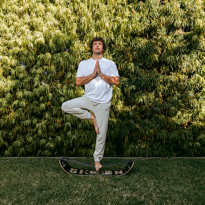 Person practicing yoga in a tree pose using a caesar slackboard plus. against a green bush background.