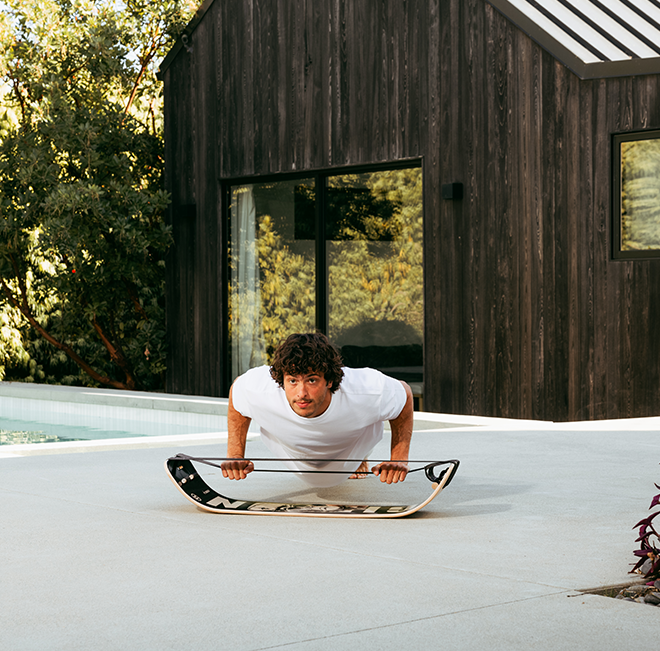 Person using a Caesar Slackboard plus to do a pushup on a patio with a wooden cabin in the background