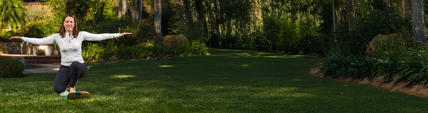 Woman balancing on a slackboard on grass in a backyard with trees and a pool in the background