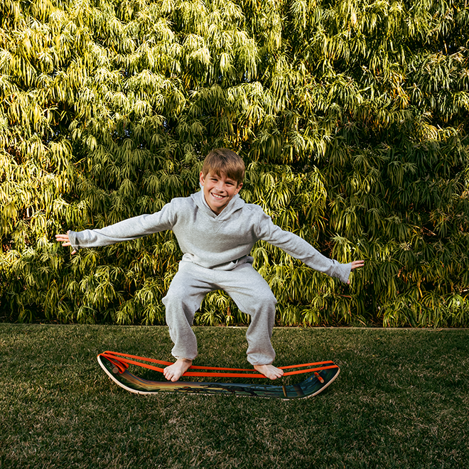 Child in a light gray outfit standing on a GIBBON Dino SlackBoard Plus with a green bushy background