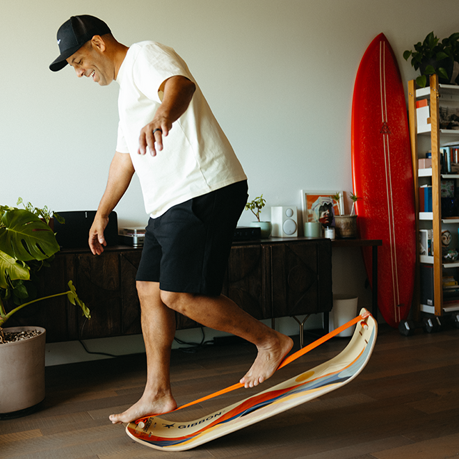 Man balancing on a sunrise slackboard in a room with a red surfboard and bookshelf.
