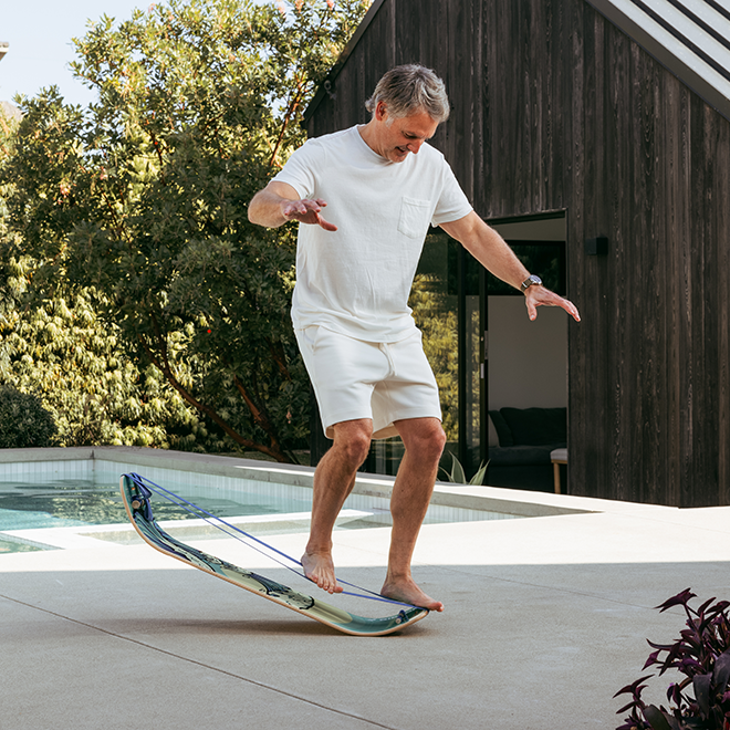 Man balancing on a waves slackboard plus poolside with a wooden building and trees in the background