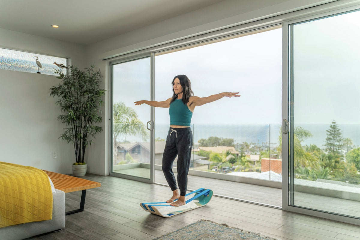 Woman balancing on a SlackBoard with arms out in a bright room with a sea view